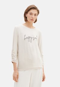 Helles beige Langarmshirt mit lockerer Passform, verziert mit gerafften Details an den Ärmeln und schwarzem gesticktem Text "happy soul" auf der Vorderseite.