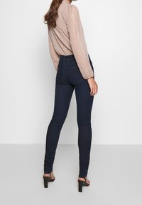High-waisted dark blue jeans with a slim fit, complemented by a sheer, light beige blouse featuring a dotted pattern and puffed sleeves.