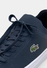 Tengerészkék kötött sneaker, lekerekített orral, texturált felülettel, fűzős dizájnnal és fehér talppal. Zöld hímzett krokodil logóval rendelkezik.