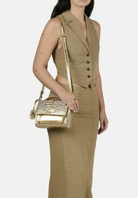 Bolso de mano dorado con textura de cocodrilo, forma estructurada, correa de hombro desmontable y detalles metálicos. Llevado con un conjunto marrón a juego.