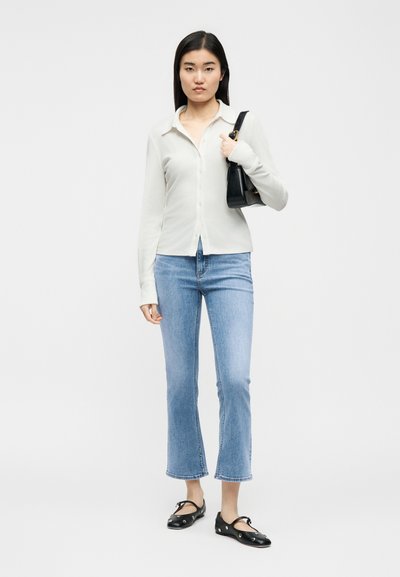 Νεαρή γυναίκα που φοράει λευκό πουκάμισο με κουμπιά, ανοιχτό μπλε cropped τζιν, μαύρες μπαλαρίνες και κρατάει μια μαύρη τσάντα ώμου.