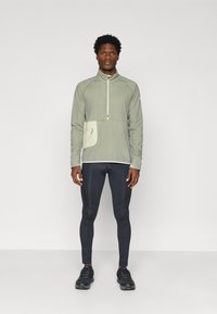 Férfi áll szemben, világoszöld negyedcipzáras pulóvert visel zsebbel, fekete, testhezálló sport leggingszel és fekete futócipővel.