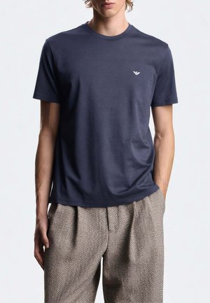 Uomo che indossa una maglietta blu navy a maniche corte con un piccolo logo bianco sul petto e pantaloni marrone chiaro con texture.