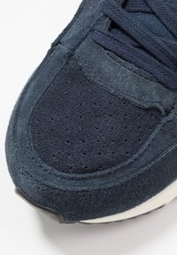 Námořnické modré semišové tenisky se strukturovaným svrškem, perforacemi a kontrastním prošíváním. Obsahují hladký špičkový kryt a gumovou podrážku.