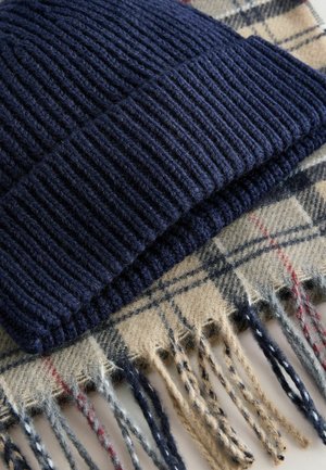 Căciulă tricotată bleumarin cu textură în dungi, asociată cu o eșarfă bej cu carouri, având margini franjurate și accente multicolore în roșu și albastru.
