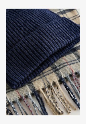 Jūras spēku adīts cepure ar ribotu tekstūru, apvienota ar bēšu krāsas ruļļoto šalli, kas rotāta ar frēzētām malām un daudzkāršu akcentu sarkanā un zilā krāsā.