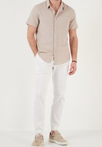 Camicia a maniche corte di colore beige chiaro con taschino sul petto, abbinata a pantaloni bianchi slim-fit e sneakers beige, su uno sfondo neutro.