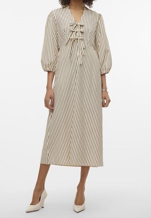 Kobieta w beżowej sukience midi z czarnymi pionowymi paskami, z bufkami z rękawami do połowy łokcia, z przodu wiązana kokardami oraz w białych butach na szpic.