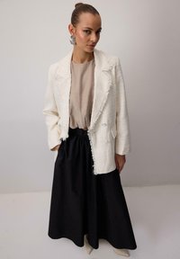 Biely tweed saká s rozstrapkanými okrajmi v kombinácii s béžovým topom a čiernou, voľnou sukňou. Obsahuje veľké strieborné náušnice a špicaté béžové topánky.