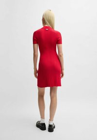 Rotes geripptes Kleid mit kurzen Ärmeln, figurbetonter Form und Rundhalsausschnitt. Enthält ein dezentes Logotag auf der Rückseite. Tragen mit klobigen schwarzen Schuhen.