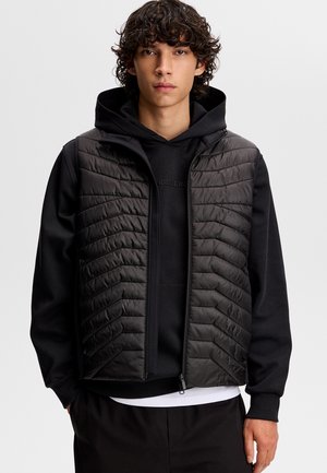 Ung mand med krøllet hår iført sort quiltet vest over sort hættetrøje og sorte bukser mod en lysegrå baggrund.