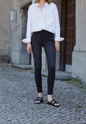 Vrouw met een wit shirt met lange mouwen, zwarte skinny jeans, zwarte sandalen, staand op een geplaveide straat vlakbij stenen gebouwen.