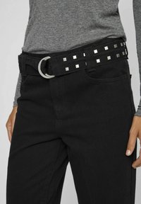 Jeans de denim negro con un amplio cinturón negro decorado con remaches metálicos cuadrados y una hebilla en forma de D plateada. La textura parece suave y resistente.