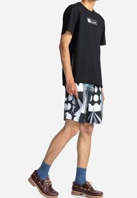 T-shirt en coton noir, short de bain à motifs dans des tons de noir, blanc et gris, chaussettes bleues et chaussures marron avec lacets et semelles en caoutchouc.