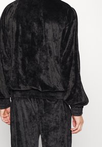 Zwarte velours trainjas met een ontspannen pasvorm. Voorzien van elastische boorden aan mouwen en zoom, een zachte textuur en een minimalistisch ontwerp.