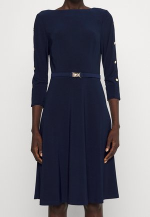 Marineblaues knielanges Kleid mit U-Boot-Ausschnitt, 3/4-Ärmeln mit goldenen Knöpfen und passendem Gürtel mit goldener Schnalle in der Taille.