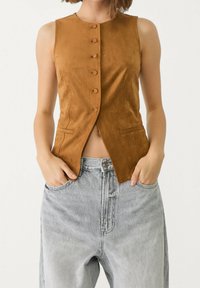 Beige suède mouwloze vest met ronde halslijn, zes knoopsluitingen en twee voorzakken, gecombineerd met lichtgrijze denim high-waisted broek.