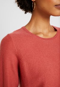 Sweter w kolorze koralowym z okrągłym dekoltem oraz strukturalnym wzorem dzianiny. Pokazuje zbliżenie na materiał i okolicę ramion.
