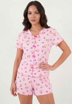 Femeie purtând un set de pijamale roz cu mâneci scurte, cu modele cu ursuleț și fundițe, având nasturi roz în formă de fundă pe partea din față a cămășii.