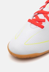 Scarpa sportiva bianca con texture rigata, dettagli giallo brillante e lacci rossi. Presenta una suola in gomma per una migliore aderenza. Primo piano dell'area della punta.
