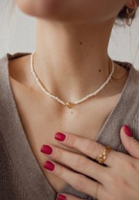 Parels choke halsketting met kleine witte parels en een gouden sluiting. Draag rond de hals boven een lichtgrijs sweater. Rode nagels zichtbaar.