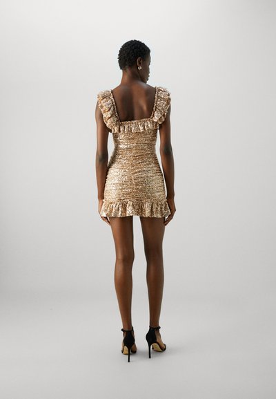 Robe courte et ajustée en or avec des bretelles volantes, tissu texturé et ourlet froncé. Portée avec des chaussures à talons hauts noires.