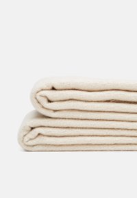 Susuktas kreminės spalvos pledas su tekstūruotu pynimu, pagamintas iš minkštos, lygios medžiagos su tvarkingomis kraštinėmis, kruopščiai sukrautas.