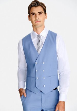 Mężczyzna w jasnoniebieskiej kamizelce dwurzędowej, dopasowanych spodniach, białej koszuli i szarym paskowanym krawacie stoi z jedną ręką w kieszeni.