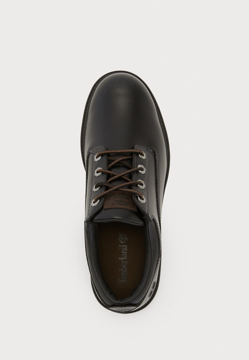 Black leather shoe with brown lace, round toe, and textured surface. Features a stitched seam and a small logo tag near the lacing.