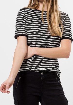 Woman with long blonde hair wearing a black and white patterned short-sleeve shirt and black pants with zippered pockets, standing against a plain background.