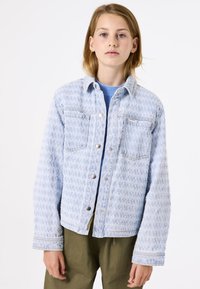 Lichtblauwe spijkerjack met een textuurpatroon, twee borstzakken en een sluiting met zilveren knopen. Draag het over een blauwe trui met ribbeldetails.