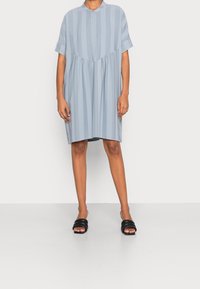 Světle modré šaty s krátkým rukávem, svislými pruhy, knoflíkovou légou, nařaseným pasem a volným střihem. V kombinaci s černými skluznými sandály.