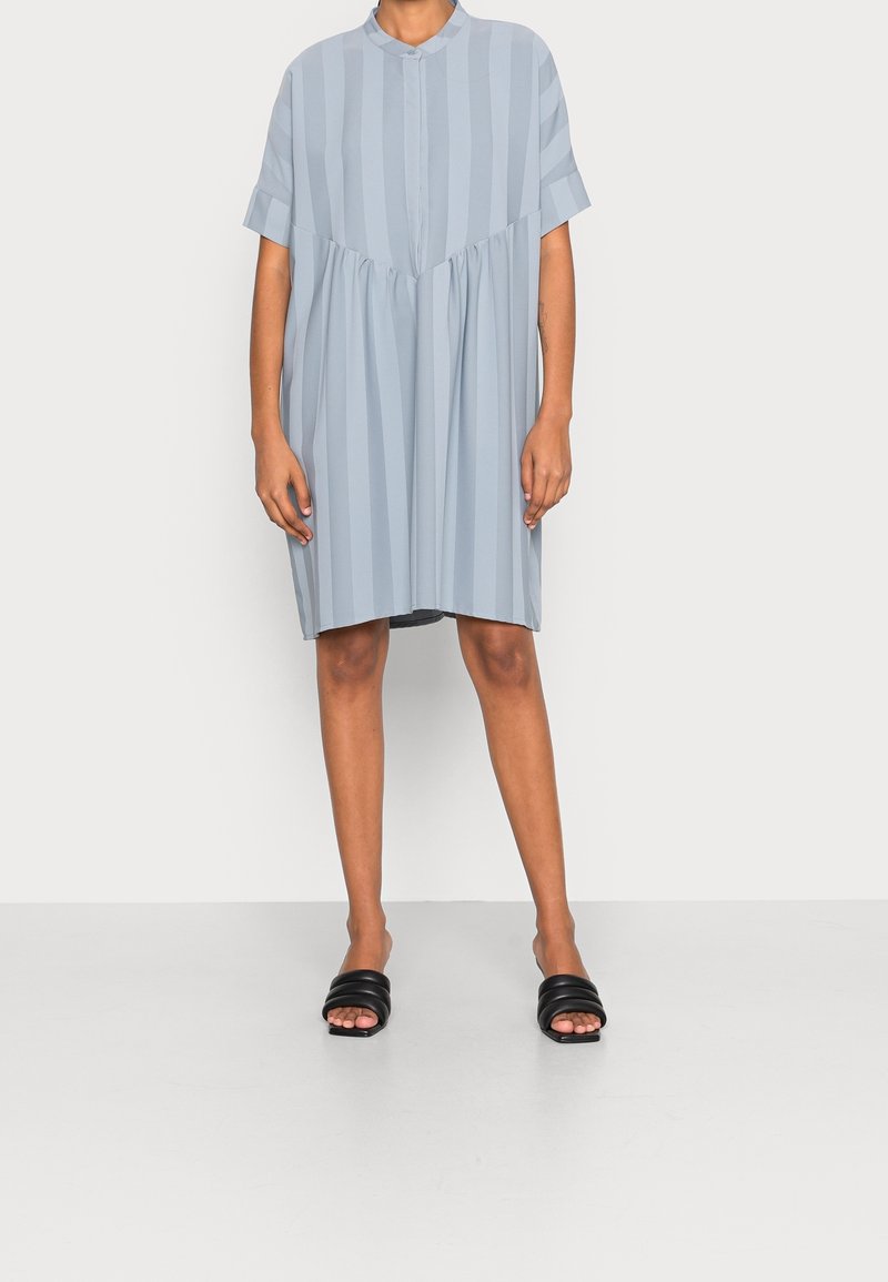 Světle modré šaty s krátkým rukávem, svislými pruhy, knoflíkovou légou, nařaseným pasem a volným střihem. V kombinaci s černými skluznými sandály.
