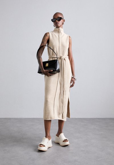 Vestito beige senza maniche con colletto alto, vita arricciata con fiocco, e spacco laterale. Borsa nera con dettagli dorati, sandali chunky con pelo bianco.