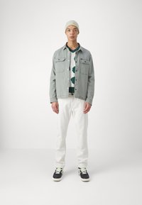 Gray denim jacket over a white and green patterned sweater, paired with white jeans and black sneakers. Model wears a light green beanie.