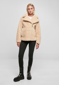 Beige nepbontjasje met een brede kraag, ritssluiting en twee voorkezen. Geselecteerd met zwarte skinny jeans en massieve zwarte laarzen.