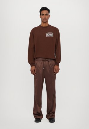 Brun langermet skjorte med hvit "Aries"-logo, kombinert med løstsittende brune satengbukser og svarte sko. Enkel og strømlinjeformet design.