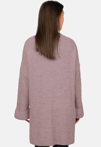 Vrouw met steil bruin haar die een losse mauve gebreide cardigan met opgerolde manchetten draagt, van achteren gezien tegen een witte achtergrond.