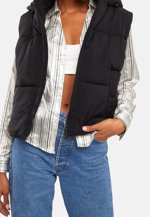 Person iført en sort polstret vest over en stribet skjorte med knapper, hvid crop top og blå jeans, med den ene hånd i lommen.
