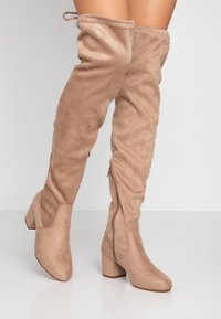 Botas de ante beige hasta la rodilla, con punta redonda y tacón cuadrado, que cuentan con una cremallera lateral y textura fruncida en la parte superior.