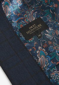 Tejido texturizado azul marino con un patrón de paisley en azul, naranja y blanco. La etiqueta dice "NEXT SIGNATURE PREMIUM TAILORING 40R."
