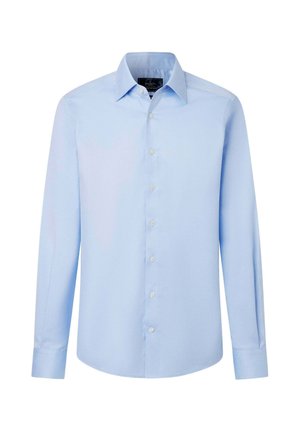 Hombre de pie vistiendo una camisa azul clara de manga larga abotonada, pantalones azul oscuro y zapatos negros contra un fondo liso y claro.