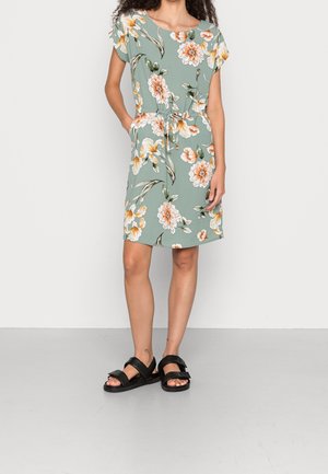 Blomstret kjole med grøn baggrund, korte ærmer, snøre i taljen og lommer. Har orange og hvide blomster, kombineret med sorte sandaler.