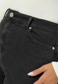 Mustat denimit housut, joissa on korkea vyötärö, metallinappikiinnitys, etupockets ja sileä pinta. Kättä, jossa on neutraali kynsilakka, lepää vyötäröllä.