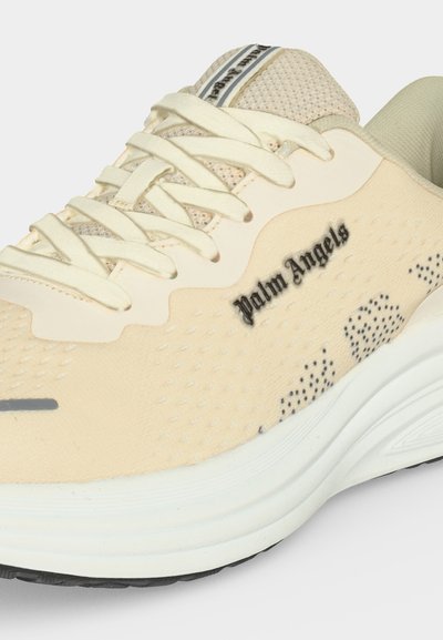 Μπεζ sneaker Palm Angels με κρεμ κορδόνια, δίχτυ στη γλώσσα, μαύρο λογότυπο με κείμενο στο πλάι και χοντρή λευκή σόλα με μαύρο πέλμα.