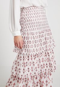 Valkoinen kuviollinen hame, jossa on kerroksellinen muotoilu ja punaisia sekä tummansinisiä timanttikuvioita. Valmistettu kevyestä kankaasta, jossa on hienoinen rakenne.