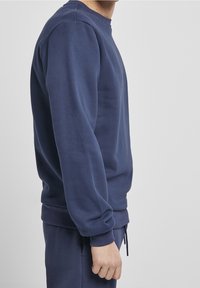 Persona vestida con un sudadera azul marino y pantalones a juego, de pie de lado contra un fondo gris claro liso.