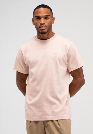 Mann steht mit hinter dem Rücken verschränkten Händen, trägt ein hellpinkes kurzärmeliges T-Shirt und beige Hose vor neutralem grauem Hintergrund.