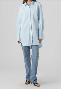 Camicia azzurra a maniche lunghe con bottoni e taschino sul petto, abbinata a jeans azzurri e infradito nere. Texture morbida, vestibilità casual.
