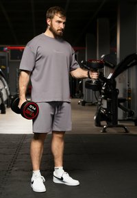 Grijs sportshirt en shorts met een gladde textuur. Witte sneakers met zwarte accenten. Houdt zwarte dumbbells vast met rode labels.